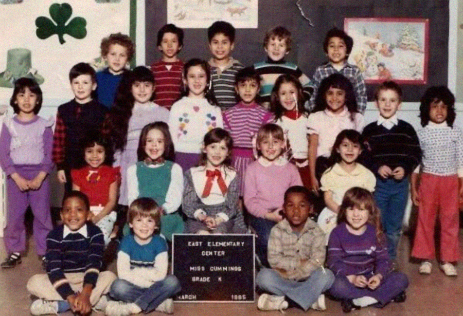 A group of young children pose for a class photo with their teacher, arranged in rows. They smile at the camera in colorful 1980s clothes. A chalkboard behind them reads “East Elementary Center, Miss Dunning, Grade K, March 1985.”