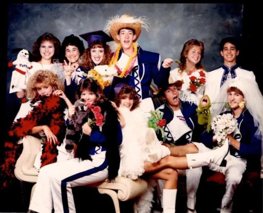 A group of young people in colorful, mismatched costumes pose playfully for a photo, some wearing hats and boas, others in athletic uniforms. They hold flowers, puppets, and stuffed animals, smiling and laughing together.
