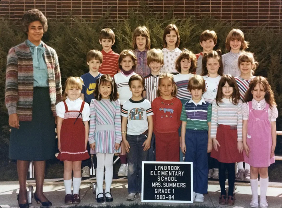 A class photo of young children and a female teacher posing outdoors in front of shrubs. A sign in front reads: “Lynbrook Elementary School, Mrs. Summers, Grade 1, 1983-84.”