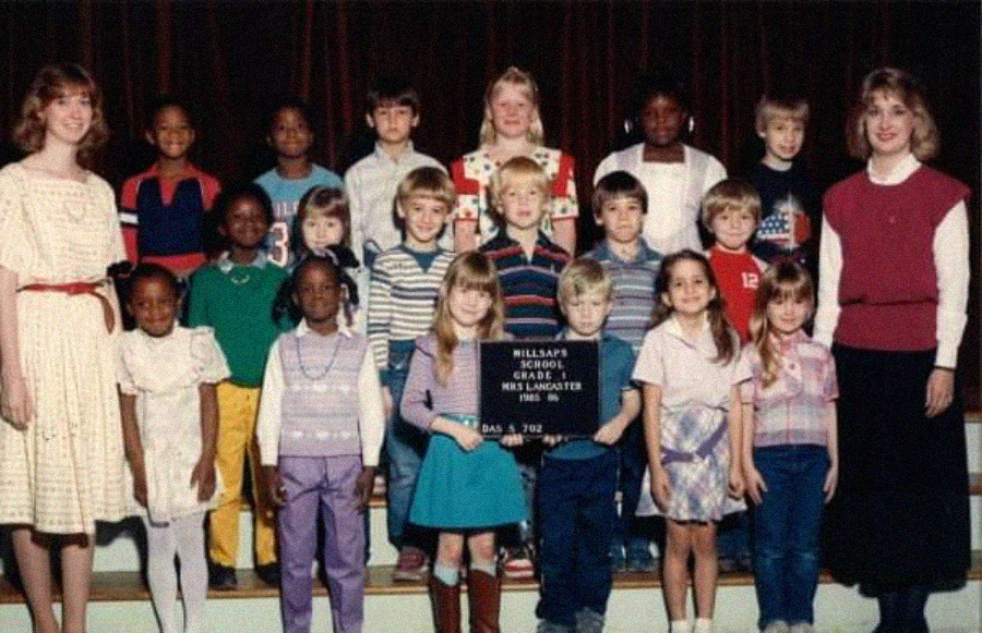 A group of school children and two adults pose for a class photo on stage. The children are arranged in three rows. A girl in the front center holds a sign with the school name, grade, teacher, and school year.