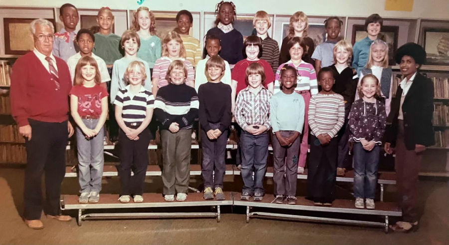 A vintage school photo shows a group of children in three rows with two adults standing at each end, all smiling, in a classroom with bookshelves in the background.