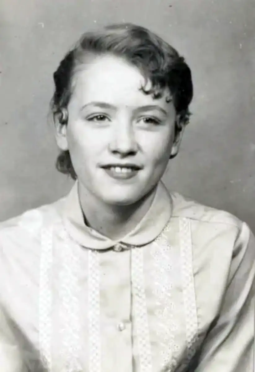 A black-and-white portrait of a young woman with light-colored hair, styled in soft waves. She is wearing a buttoned shirt with lace detailing and is looking slightly to the side, smiling gently.