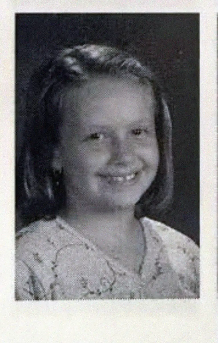A black and white yearbook-style portrait of a young girl with straight, shoulder-length hair, smiling and wearing a patterned top. The background is plain and dark.