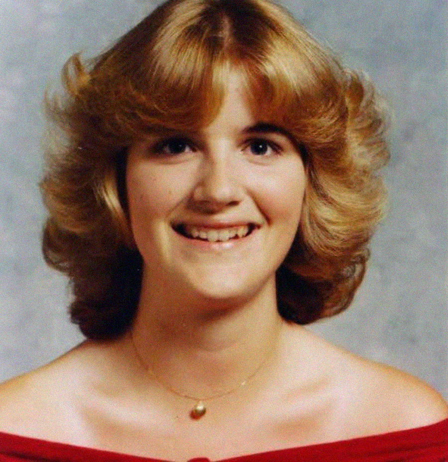 A young woman with feathered, shoulder-length blonde hair smiles at the camera. She wears an off-the-shoulder red top and a gold necklace with a pendant, posed in front of a mottled gray background.