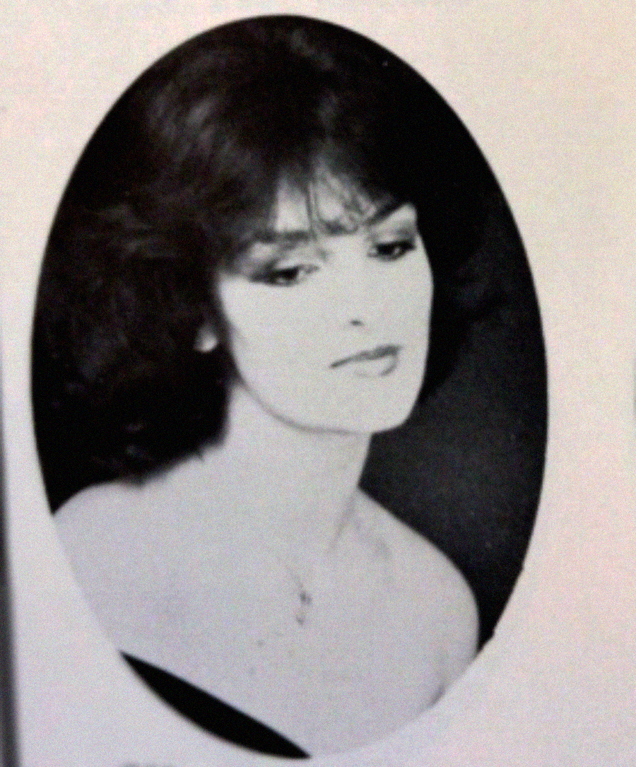 Black-and-white portrait of a woman with dark, wavy hair and bangs, looking slightly down and to the side. She wears an off-the-shoulder top and a delicate necklace, set against a dark background inside an oval frame.