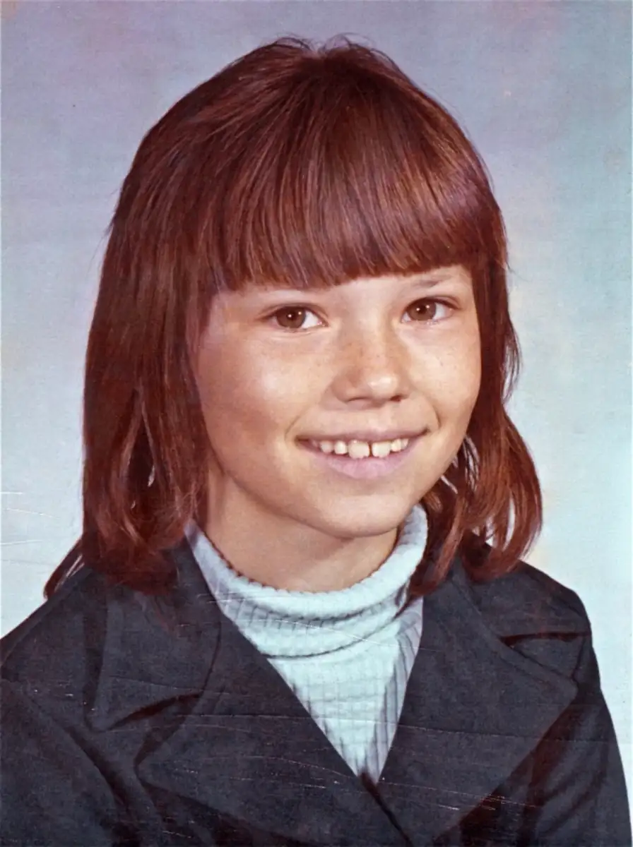 A young girl with straight brown hair and bangs smiles at the camera. She is wearing a dark jacket over a light turtleneck sweater, and the background is a plain, light color, typical of a school portrait.