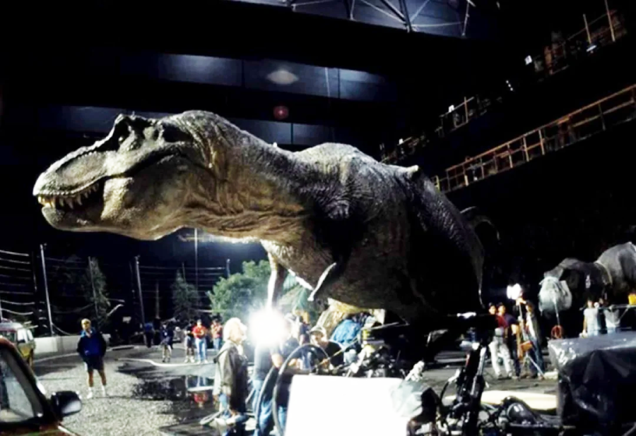 A large T. rex animatronic model on a film set, surrounded by crew members and equipment, with bright lights illuminating the scene inside a soundstage.