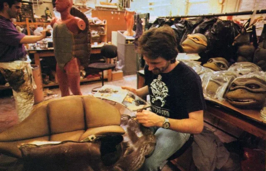 A man works on a turtle costume piece in a workshop filled with masks and costume parts. Another person stands nearby next to a mannequin wearing a muscle suit. The room is cluttered with materials and tools.