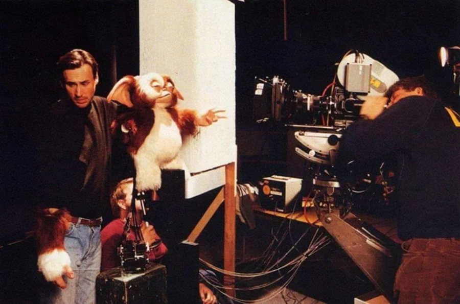 Two people operate a large puppet of Gizmo from the film "Gremlins" on a set, with one person controlling the puppet and another working behind a professional movie camera.