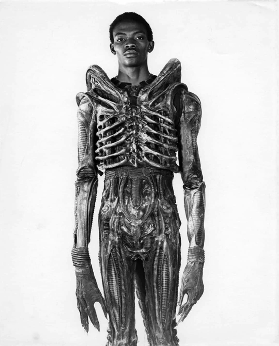 A person stands wearing a detailed, sculpted alien costume with rib-like chest armor, elongated arms, and intricate textures, against a plain white background.