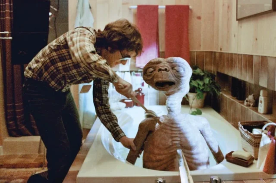 A person in a plaid shirt uses a washcloth to clean a friendly alien sitting in a bathtub inside a rustic bathroom with brown tiles and wooden walls.