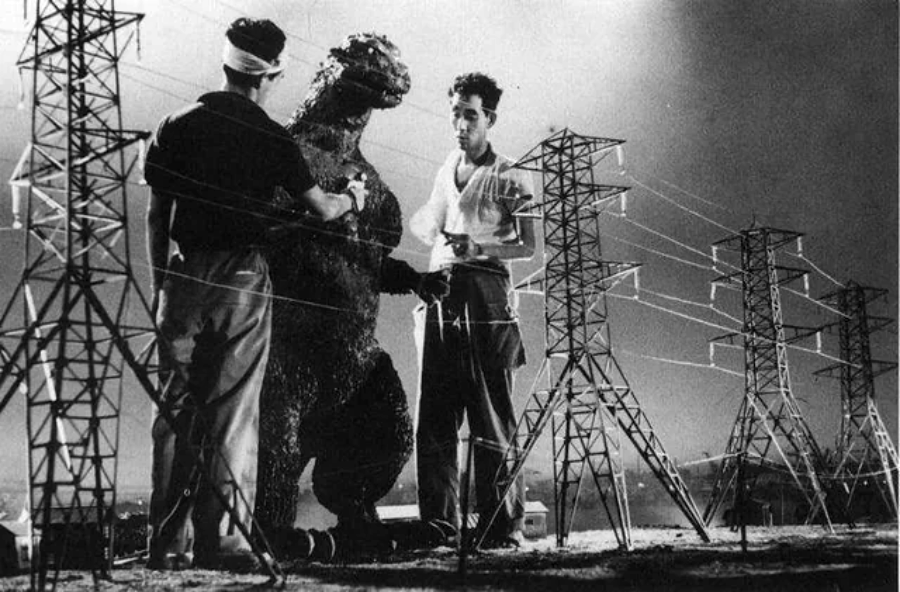Two men stand next to a person in a Godzilla suit on a miniature set with model power lines and towers, likely behind the scenes of an old Godzilla movie production.