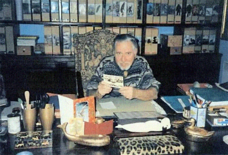 A man with a beard sits at a cluttered desk, holding a photograph. Behind him, shelves are filled with files and books. The desk is covered with papers, a pen holder, framed photos, and various objects.