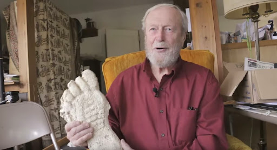 An older man with a white beard and red shirt sits in a room, smiling and holding a large plaster cast of an oversized foot. Boxes, papers, and furniture are visible in the background.