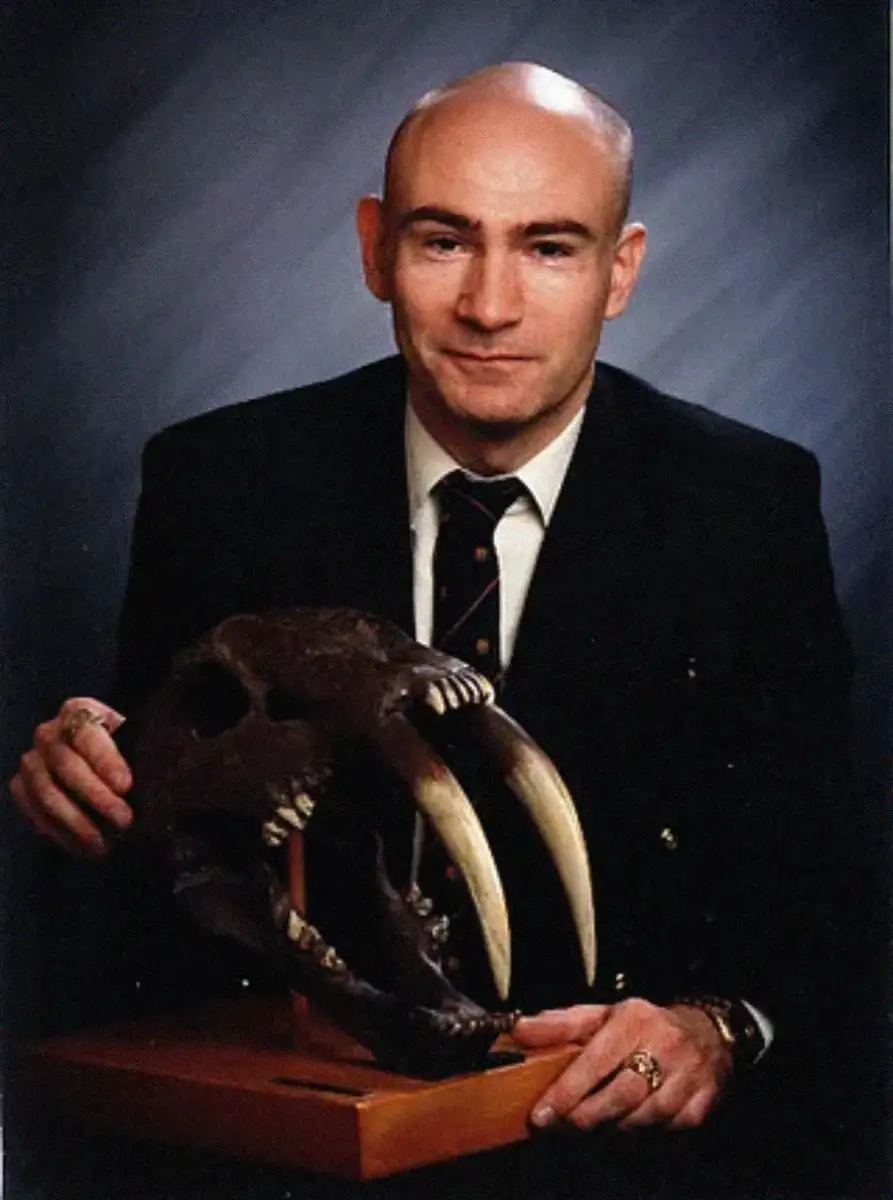 A man in a suit poses for a portrait while holding a large saber-toothed cat skull replica with long, curved canine teeth, set on a wooden base.