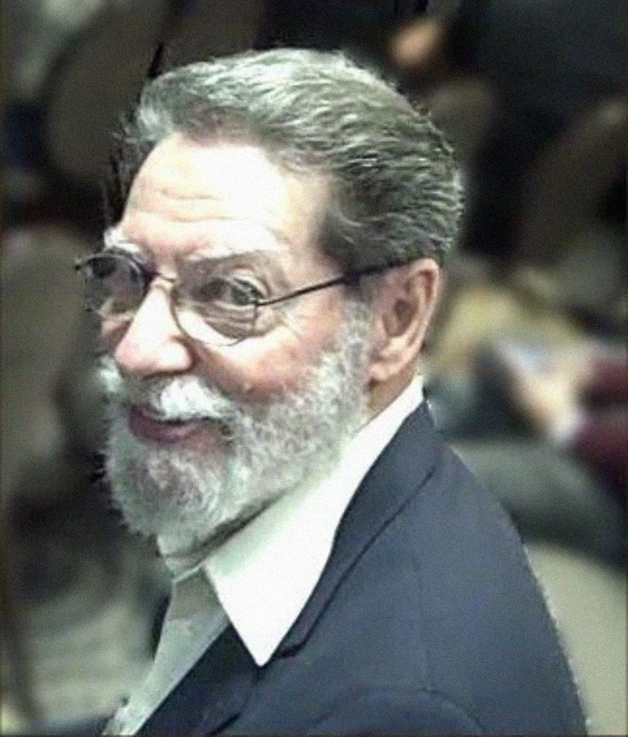 An older man with glasses, gray hair, and a beard smiles while looking over his shoulder. He is wearing a dark suit jacket and a light-colored shirt, seated in a room with blurred people in the background.