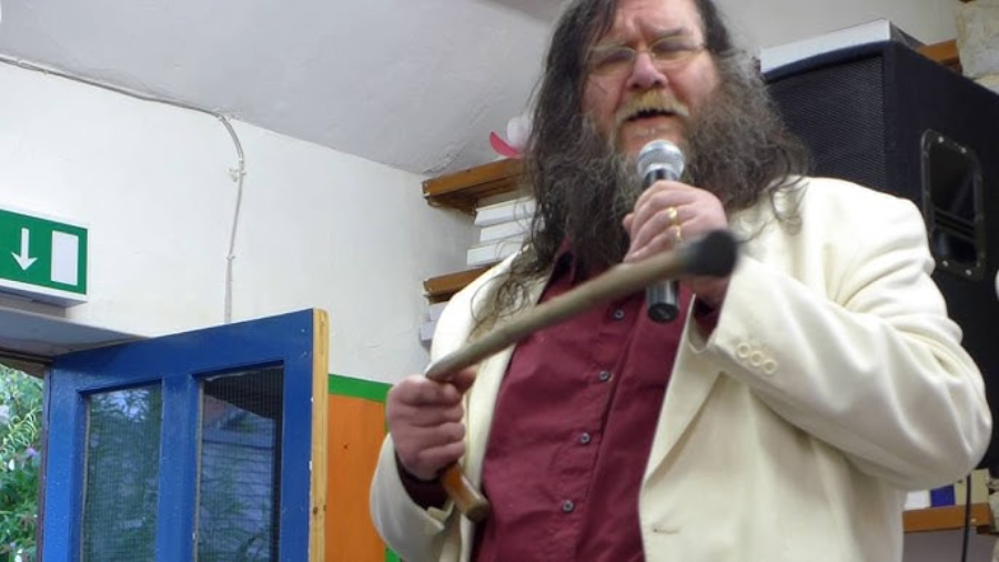 A man with long hair and a beard, wearing glasses, a cream-colored blazer, and a maroon shirt, speaks into a microphone while holding a wooden cane indoors near a blue door and shelves.