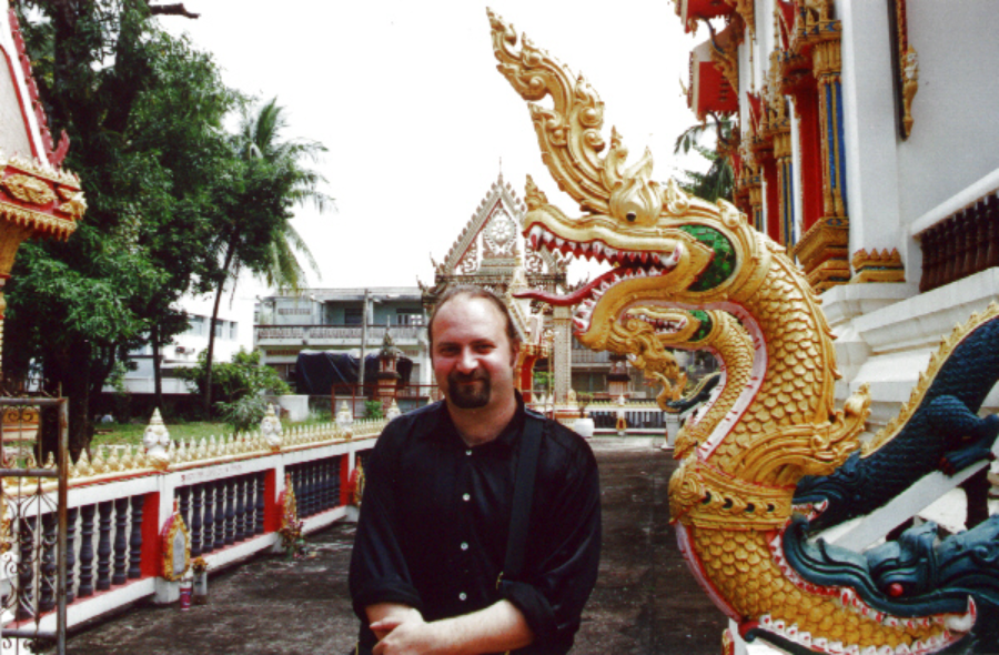 A man stands smiling in front of an ornate golden dragon statue at a colorful temple, with palm trees and buildings visible in the background.