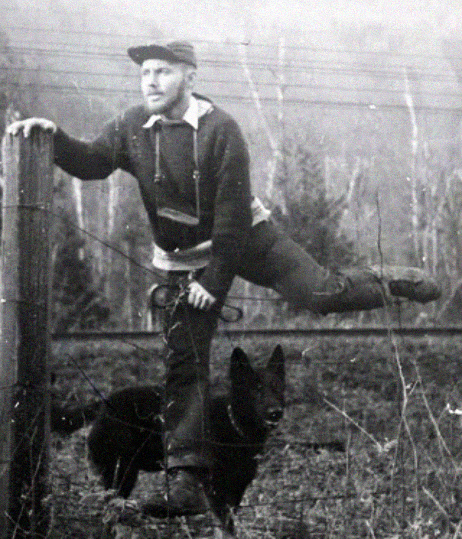A man climbs over a wooden fence, resting one leg on the top rail while holding onto a post. A dark-colored dog stands beside him, partially obscured, in a rural outdoor setting with trees in the background.