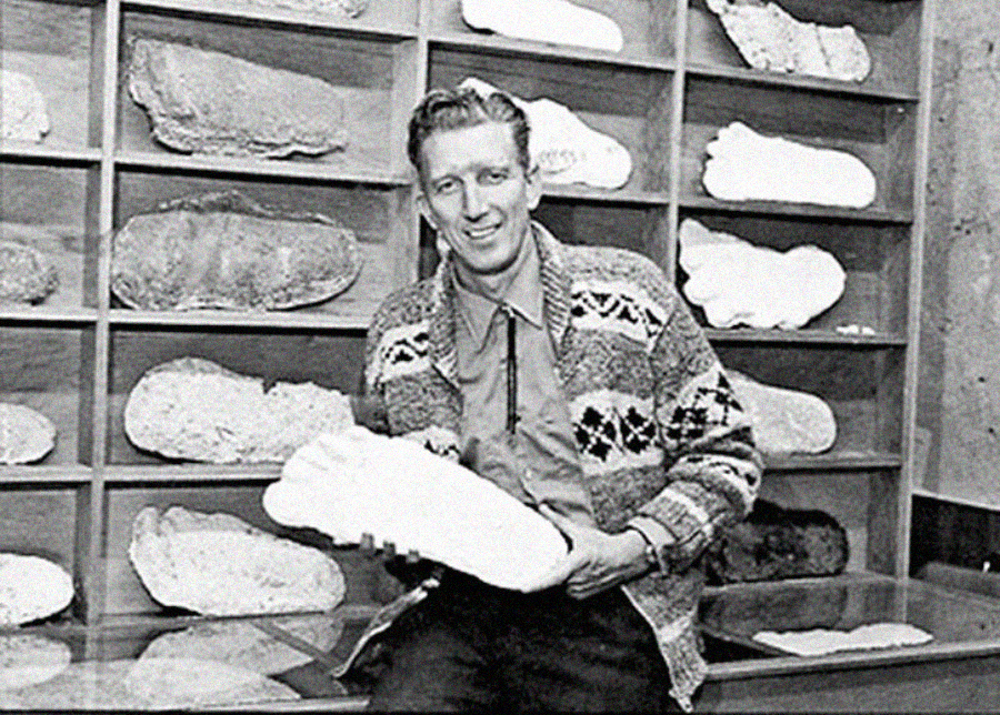 A man sits in front of shelves filled with large plaster casts of footprints, holding one cast in his hands and smiling at the camera.