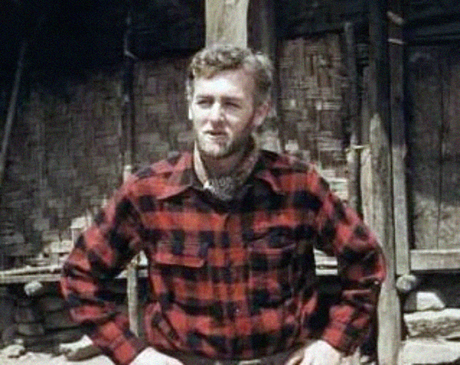 A man with a beard and short hair stands outdoors wearing a red and black plaid shirt. He has his hands on his hips and appears to be in a rustic setting with wooden structures and bamboo walls behind him.