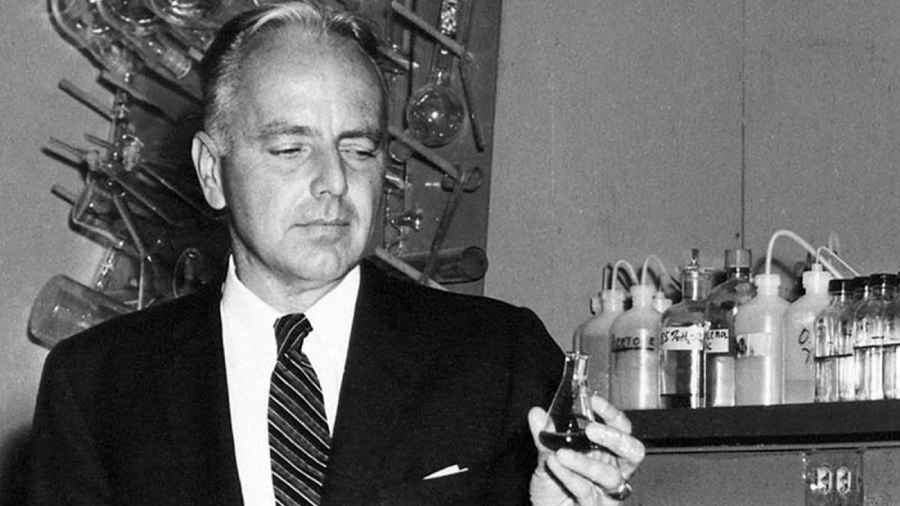 A man in a suit and tie holds a small laboratory flask with liquid, standing in a lab with shelves of glass bottles and scientific equipment in the background.