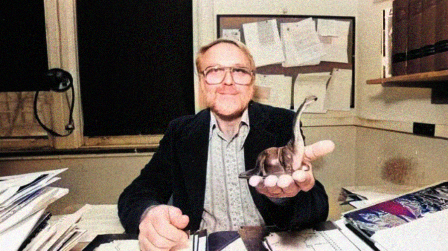 A man with glasses sits at a desk in an office, smiling and holding a small model of a sauropod dinosaur in his hand. Papers, books, and files are scattered around him, and a bulletin board is visible behind.