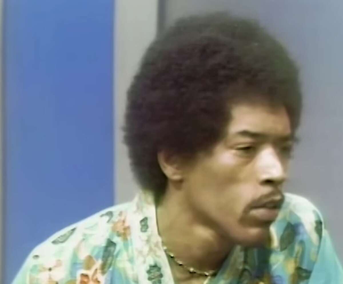A man with an afro hairstyle and a thin mustache wears a floral shirt and a beaded necklace, looking thoughtful in a studio setting with blue and gray background panels.