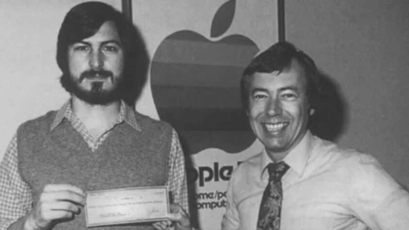 Two men stand smiling in front of a vintage Apple Computer logo; one holds a check while both face the camera in this black-and-white photograph.