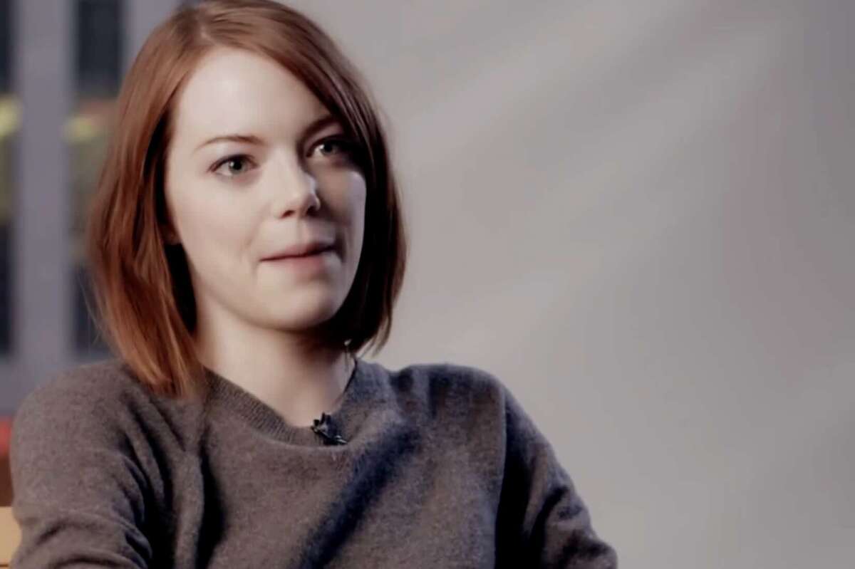 A young woman with shoulder-length reddish-brown hair, wearing a grey sweater, sits and looks slightly to the side with a soft smile against a blurred background.