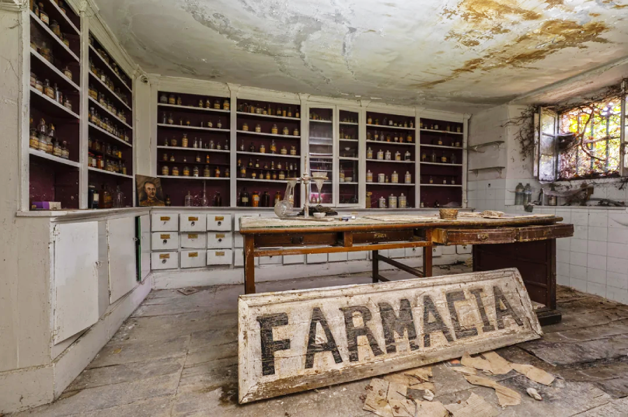 An abandoned pharmacy with dusty shelves full of old bottles, a weathered wooden counter, and a large broken “FARMACIA” sign lying on the dirty floor. Light filters in through a stained-glass window.
