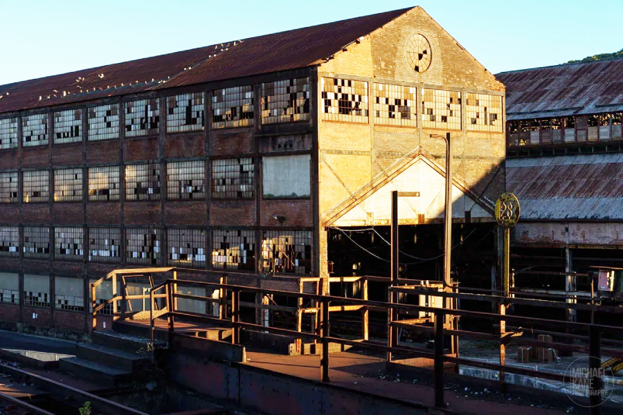 An old, abandoned industrial building with rusted metal roof, broken windows, and weathered brick walls, bathed in late afternoon sunlight. Foreground shows metal railings and industrial debris.