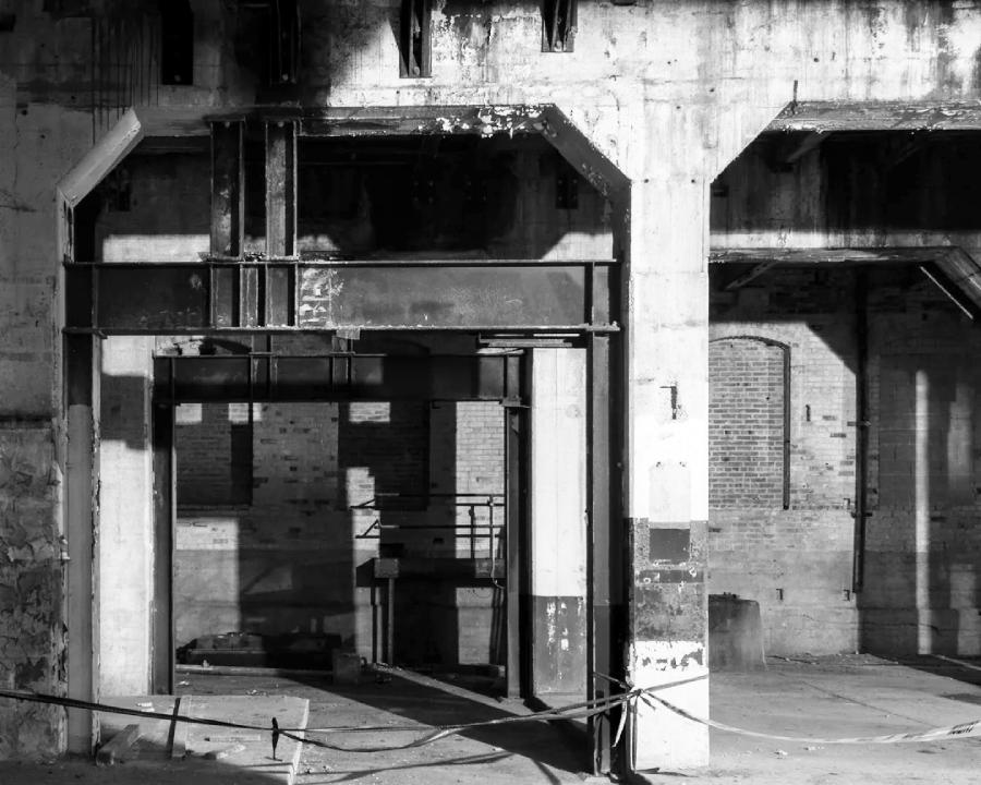 Black and white photo of an abandoned industrial building interior with exposed concrete, brick walls, metal beams, and deep shadows. A rope barrier stretches across the foreground.