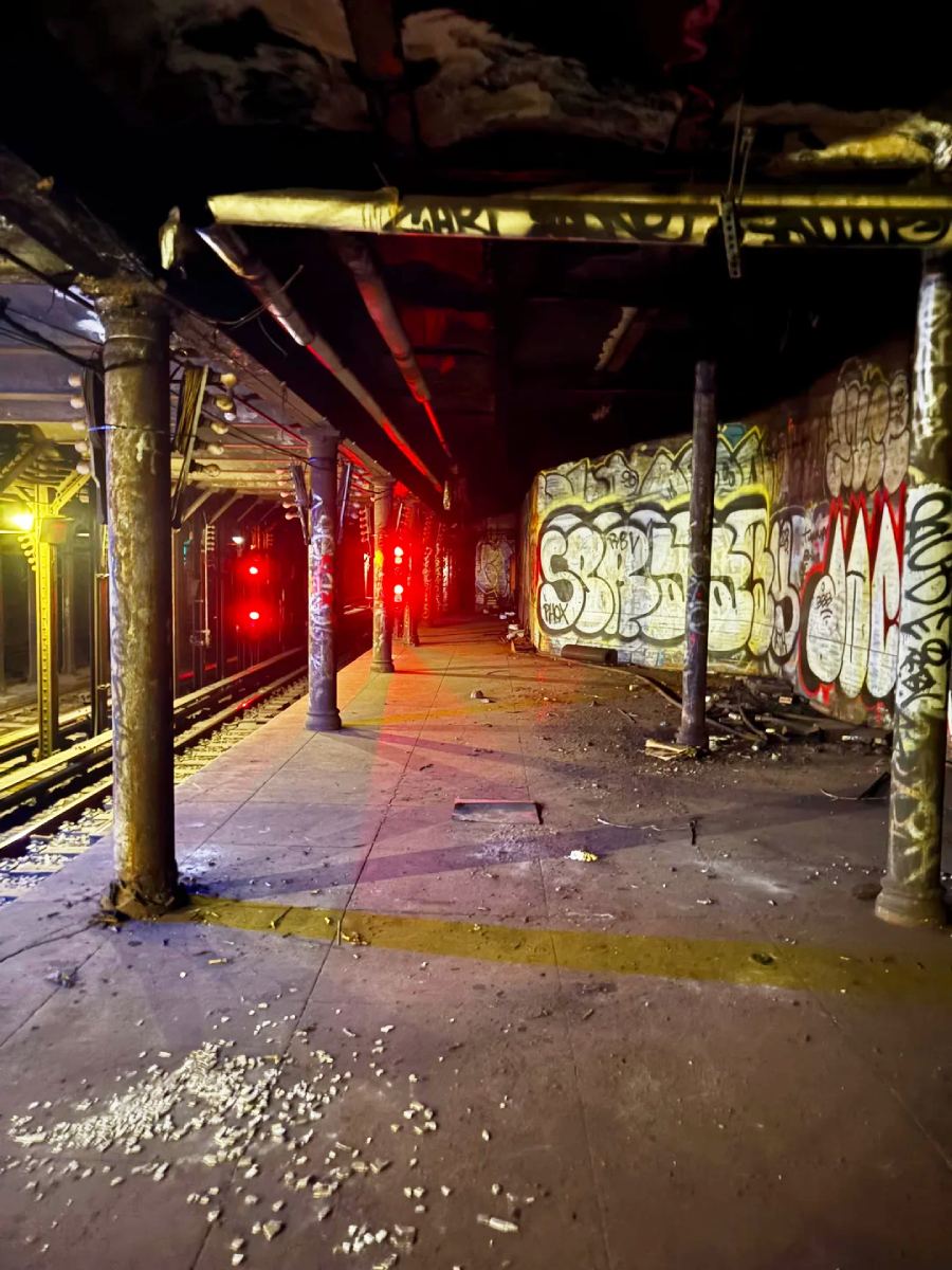 An abandoned subway platform with graffiti-covered walls, broken columns, debris on the ground, and red signal lights illuminating the otherwise dark station.