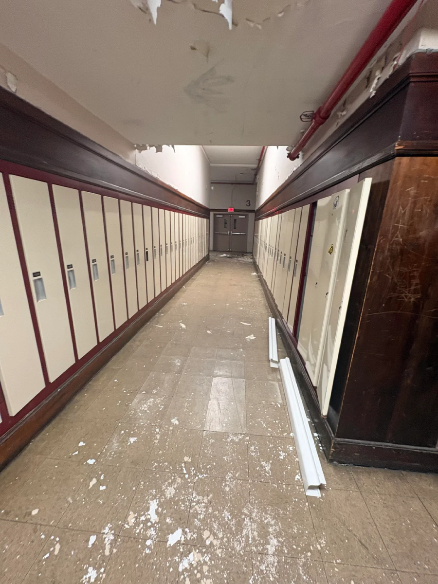 A long hallway in a school with beige lockers on both sides, peeling paint on the ceiling, and debris scattered on the floor. The hallway leads to double doors at the end.