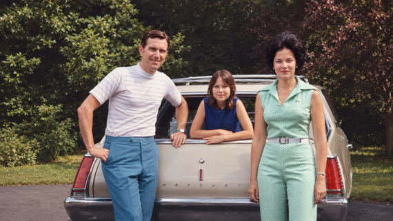 A man, a woman, and a young girl pose in front of a vintage Mercury station wagon on a driveway, with green trees and grass in the background.