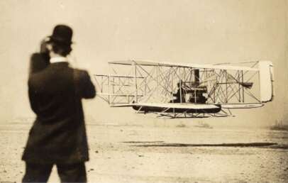 A black-and-white photo shows a man in a suit and bowler hat watching an early biplane, likely the Wright Flyer, taking off or landing on a flat, sandy terrain.