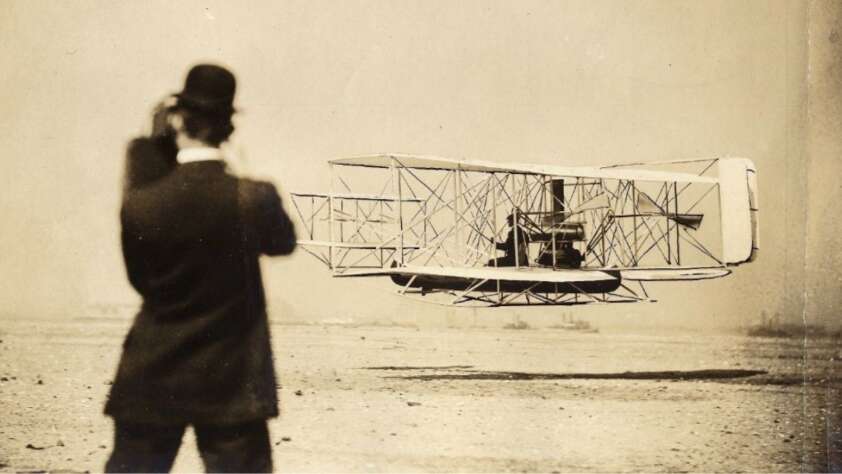 A black-and-white photo shows a man in a suit and bowler hat watching an early biplane, likely the Wright Flyer, taking off or landing on a flat, sandy terrain.