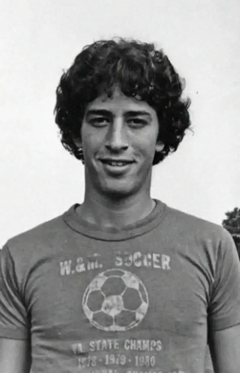 Young man with curly hair smiles at the camera, wearing a "W.M. Soccer State Champs 1978/1979/1980" T-shirt with a soccer ball graphic. The image is in black and white.