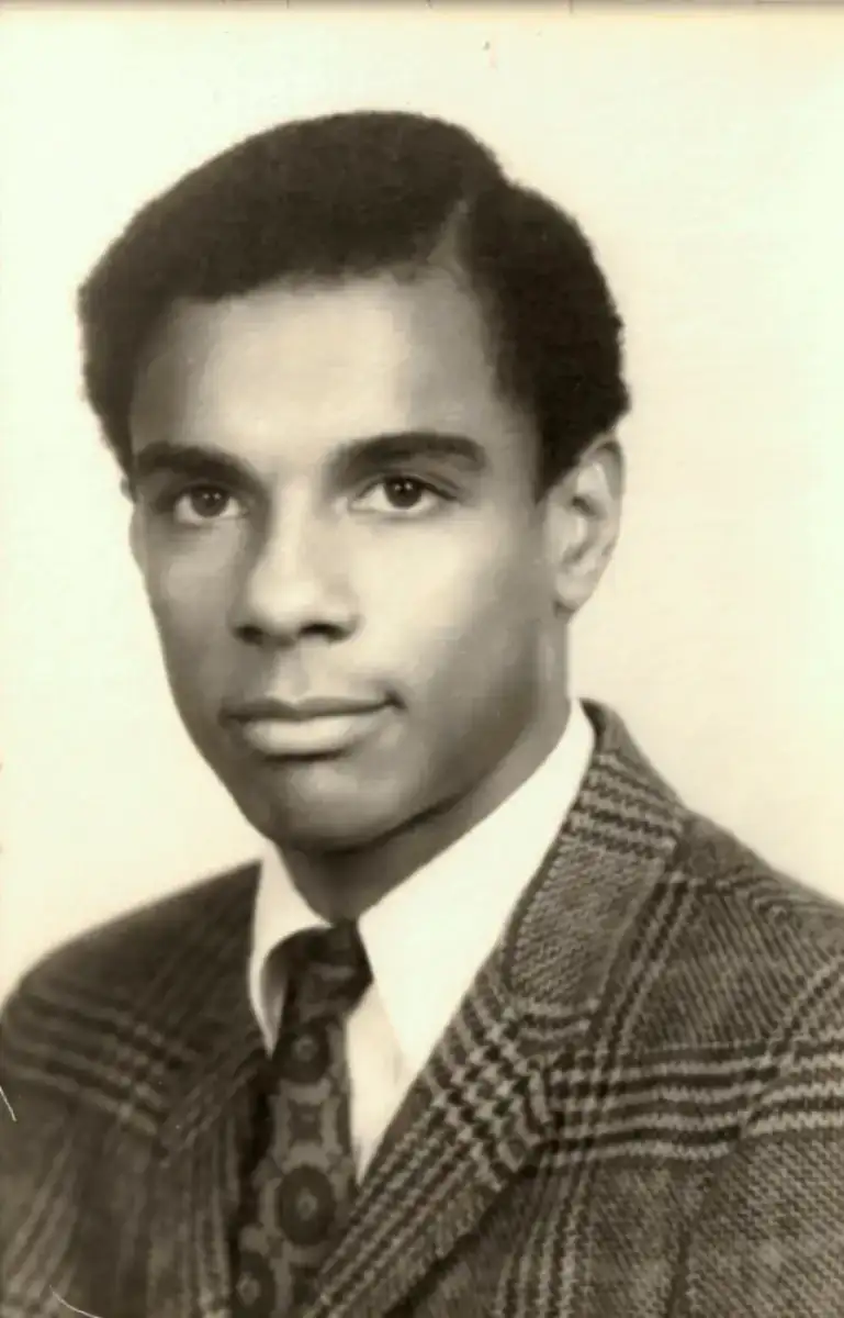 A young man with short, neatly styled hair wearing a plaid suit jacket, white shirt, and patterned tie, poses for a formal black-and-white portrait against a plain background.