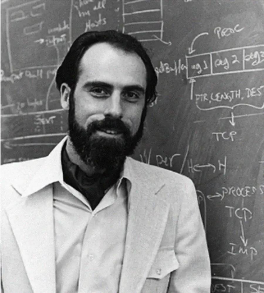 A man with a beard and dark hair stands in front of a chalkboard filled with diagrams and technical notes, wearing a light-colored suit jacket and open-collared shirt.
