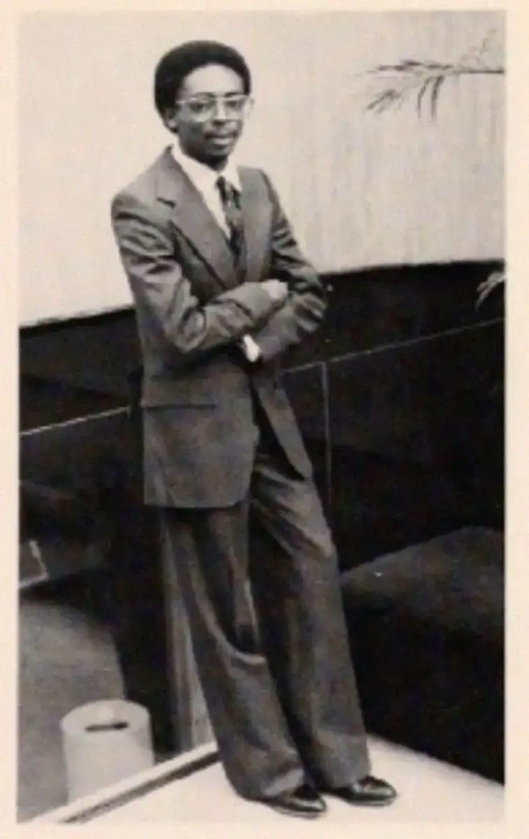 A man in a suit and tie stands indoors with arms crossed, leaning against a glass partition. He wears glasses and dress shoes; the setting includes modern furniture and a potted plant in the background. The image is in black and white.