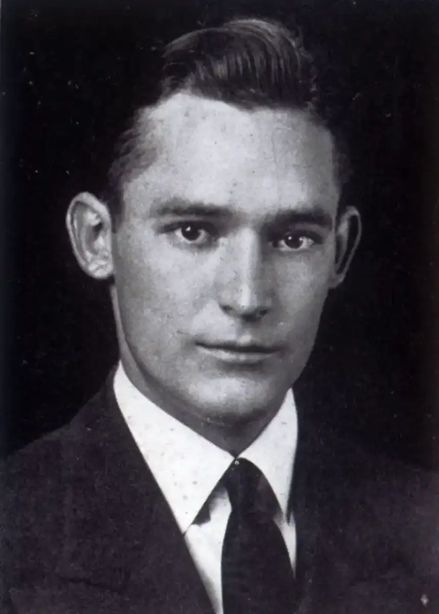 A black and white portrait of a young man with short, neatly combed hair, wearing a dark suit, white shirt, and dark tie, looking directly at the camera with a neutral expression.