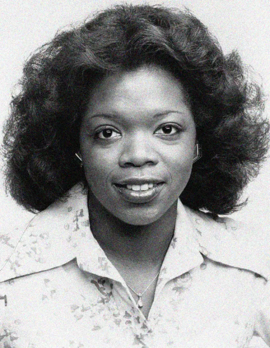 A young woman with medium-length, curly hair smiles at the camera. She is wearing a light-colored, collared shirt with a subtle floral pattern and a small necklace. The photo is black and white.