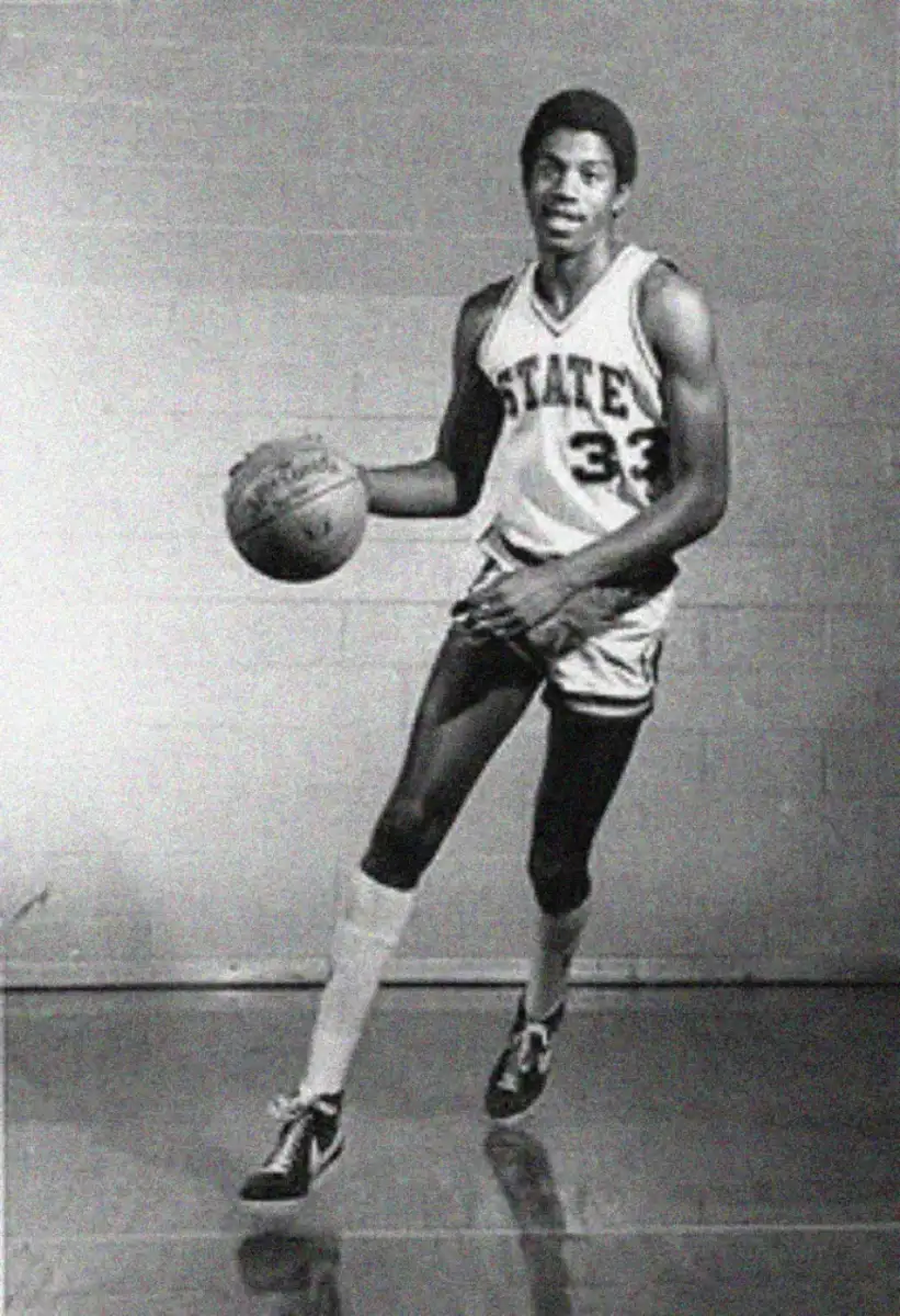 A young male basketball player in a "STATE" jersey with the number 33 dribbles a basketball. He stands on a gym floor against a plain wall, wearing high socks and sneakers. The image is black and white.
