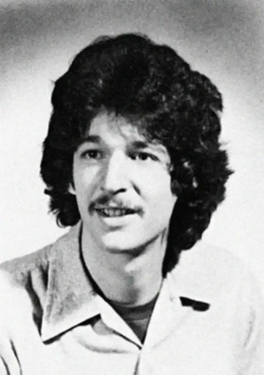 Black and white portrait of a young man with thick, curly hair and a mustache, wearing a collared shirt and looking slightly to the side, smiling softly.