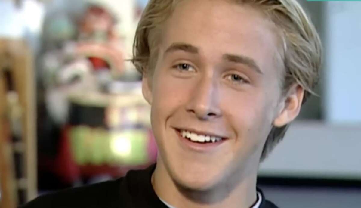 A young man with blonde hair and fair skin is smiling warmly. He is wearing a black shirt, and the background is blurred with pastel colors.