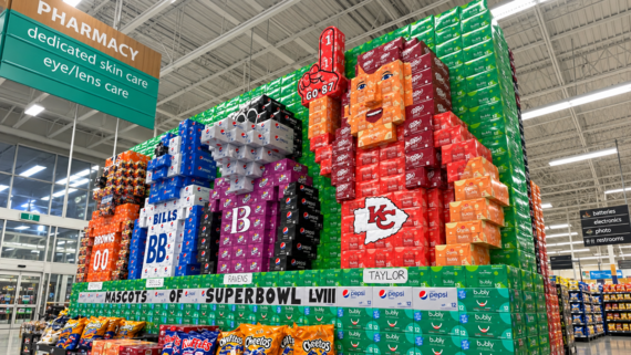 Large soda box display in a store forms mascots of NFL teams, including the Kansas City Chiefs, with a “Go 87!” sign, next to pharmacy and snacks aisles.