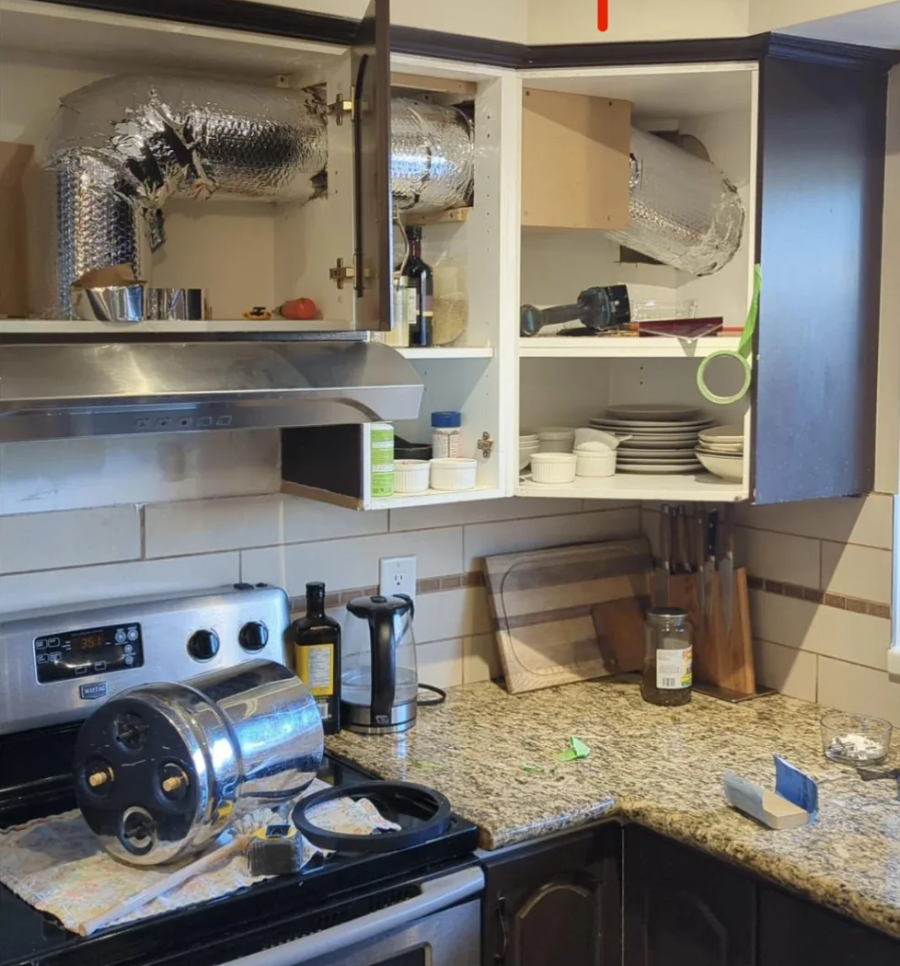 A kitchen with open cabinets revealing exposed ductwork inside. Dishes and kitchen items are partially displaced, with appliances, bottles, and a tipped-over pressure cooker on the countertop, giving a cluttered appearance.