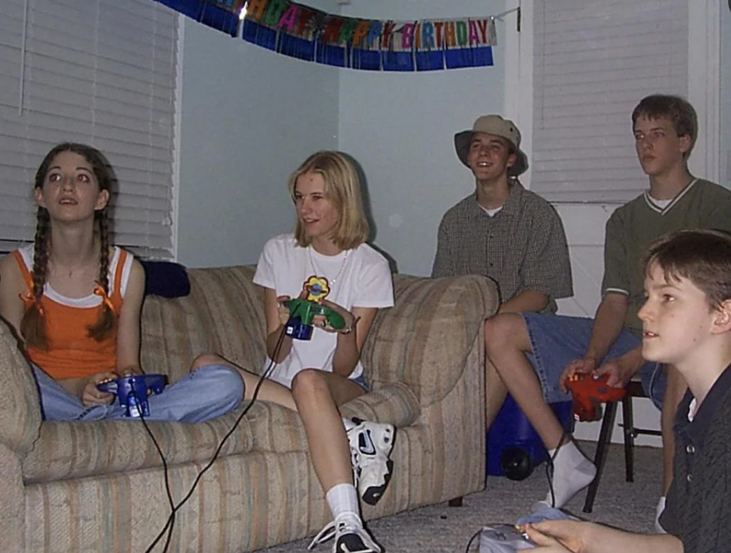 Five teenagers are sitting in a living room, playing video games together. Two are on a couch and three are on the floor. A "Happy Birthday" banner hangs on the wall behind them. Everyone looks engaged and happy.